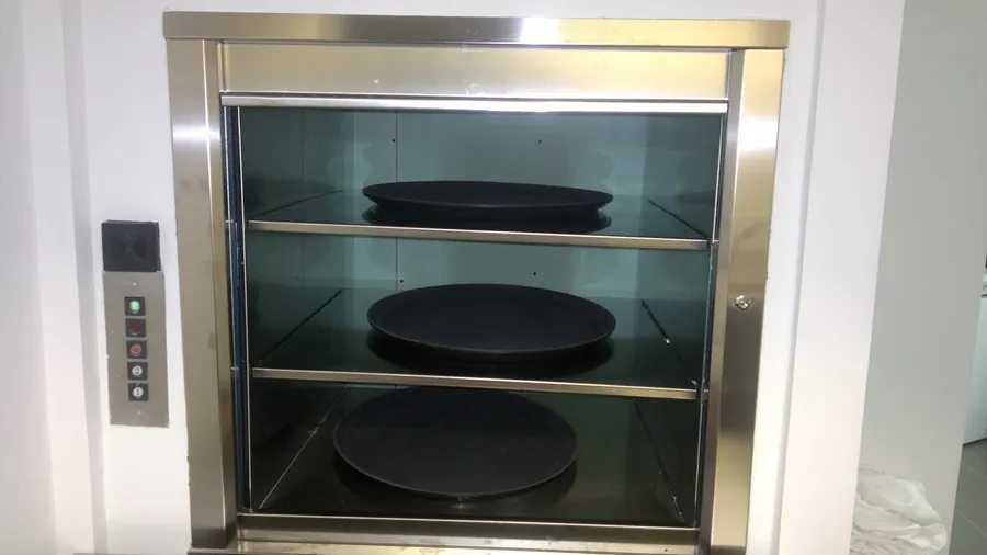 Montaplatos de acero inoxidable instalado por Plataformas y Equipos de Elevación, con la puerta abierta y bandejas de servicio en sus tres estantes